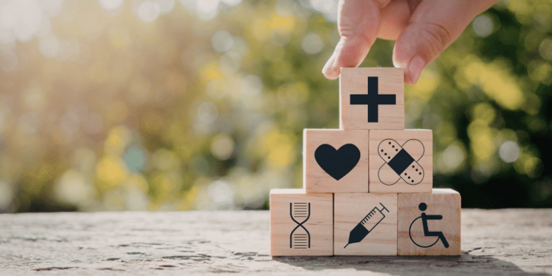 A person stacks blocks on a rock in a park on a sunny day. The blocks have healthcare symbols on them.